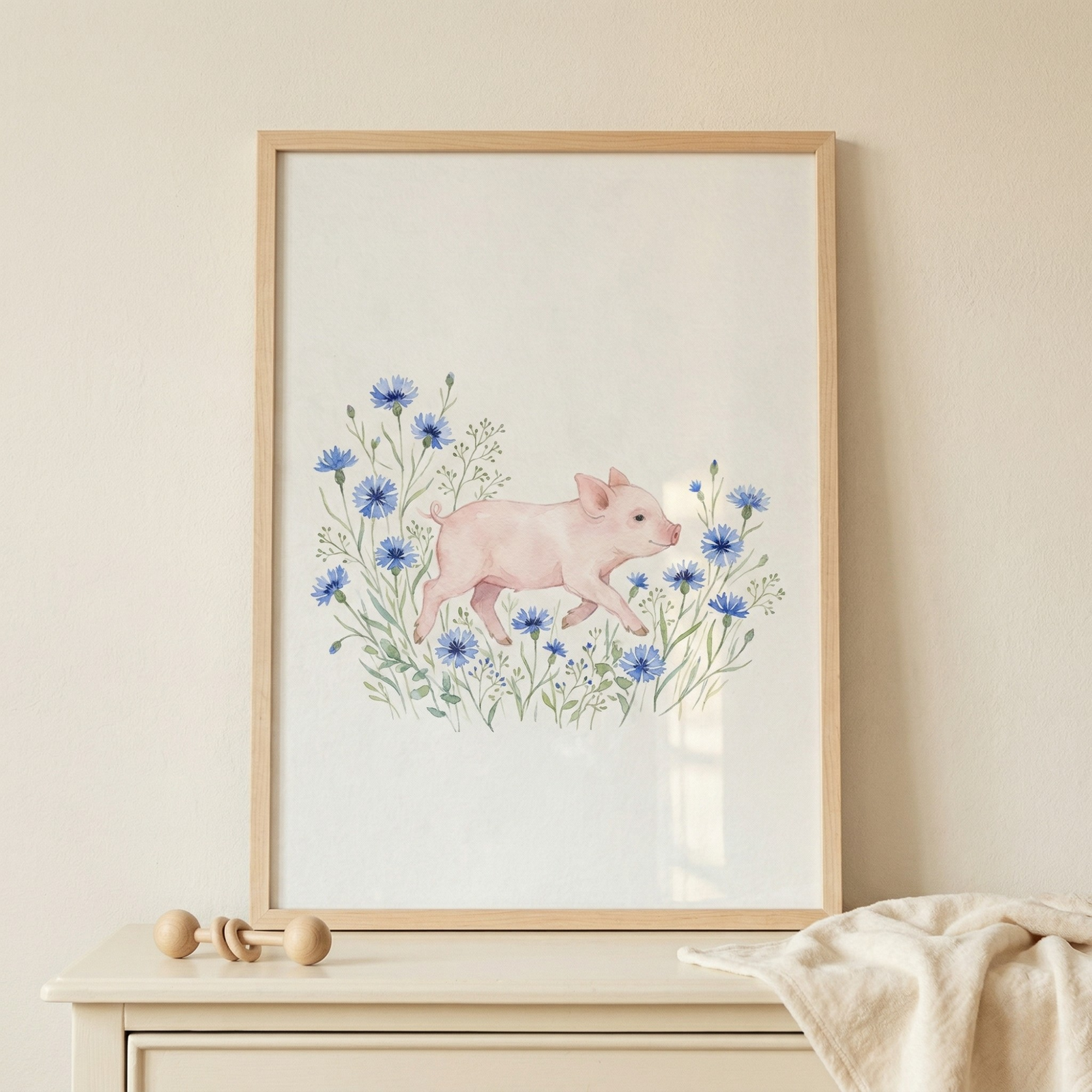 A soft watercolor-style illustration of a small pink piglet trotting through a cluster of delicate blue wildflowers. The artwork is centered on a white background and encased in a light wood frame. It sits on a beige surface alongside a small wooden rattle and a soft cream-colored blanket, set against a plain neutral wall in a minimalist nursery setting.