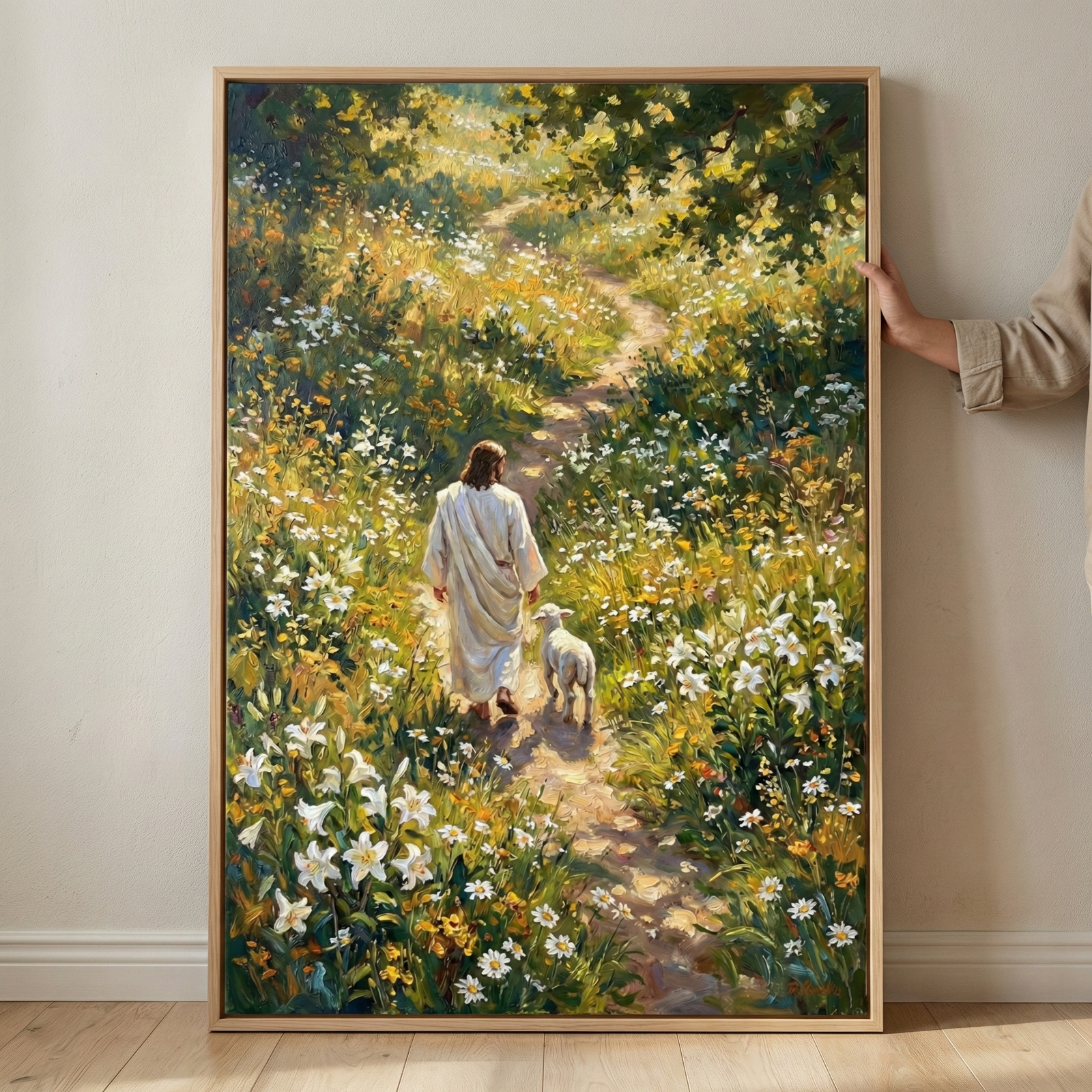 A large framed vertical artwork shows Jesus Christ from behind, walking down a sun-dappled dirt path in a long white robe. A small white lamb walks faithfully by His side. The path winds through a garden of blooming white lilies and daisies. Warm sunlight filters through leafy trees overhead, creating a peaceful scene. The style uses thick, visible brushstrokes. A hand holds the light wood frame against a plain wall above a light-colored wooden floor.