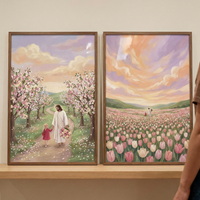 Two framed vertical illustrations rest on a light wood shelf. The left piece depicts Jesus in a white robe walking hand-in-hand with a young girl in a red dress down a dirt path. They are surrounded by flowering pink trees, and Jesus carries a bouquet. The right piece shows the same pair as small figures in the distance of a vast field of pink and white tulips. Above them, a dramatic, swirling sky is filled with pastel shades of violet, orange, and yellow.