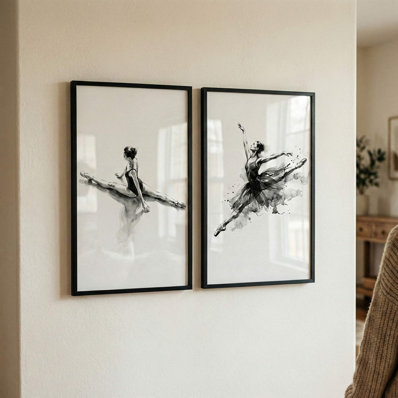 Two framed monochromatic artworks hang side-by-side on a light-colored wall. The artwork on the left features a ballet dancer in a dark leotard, depicted in a mid-air split. The artwork on the right shows a dancer in a dark tutu captured in a dynamic, expressive leap with one arm raised. Both pieces are rendered in a minimalist style with ink-like washes and splatters. They are encased in thin, black rectangular frames. A glimpse of a home interior is visible in the background to the right.