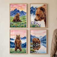 Four framed watercolor illustrations of brown bears are displayed in a 2x2 grid on a light wall. Top-left shows a bear sitting in wildflowers with blue mountains under a pink sunset. Top-right is a close-up of a bear's head with purple flowers. Bottom-left features a bear standing upright in a meadow. Bottom-right captures a bear drinking from a lake with its reflection. Each artwork has a light wood frame. A person in a beige sweater is partially visible on the right.