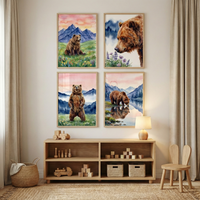 A set of four watercolor-style illustrations featuring brown bears is displayed on a light-colored wall. Each artwork is set in a simple wooden frame and arranged in a grid pattern. The scenes depict bears in various natural settings: one sits peacefully in a flowered meadow, another is shown in a close-up portrait, a third stands tall in a grassy field, and the last drinks from a mountain lake. All pieces share a soft color palette of blues, pinks, and earth tones.

