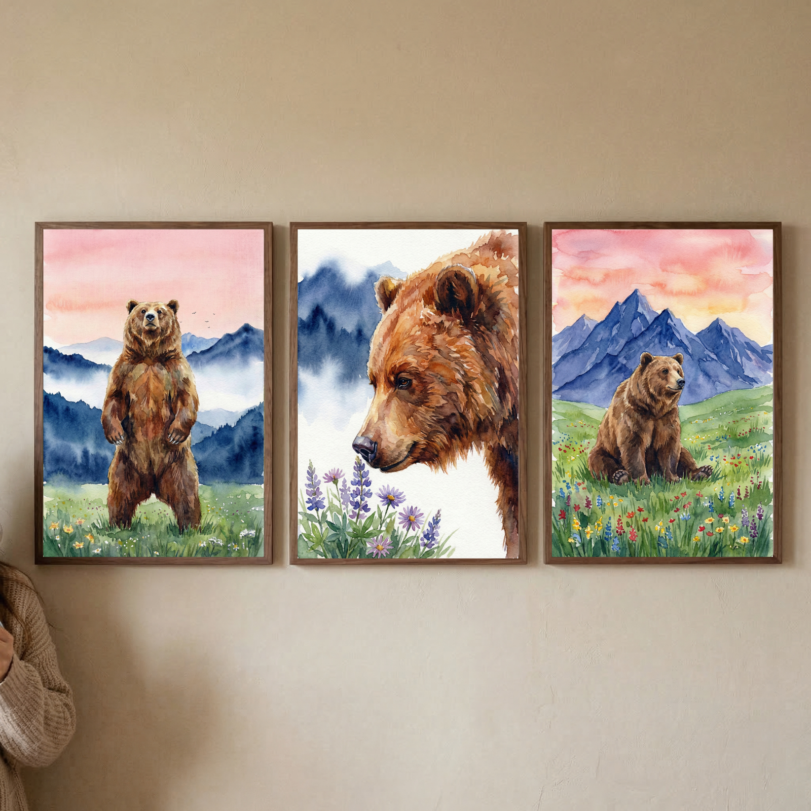 Three framed watercolor illustrations hang in a row on a plain wall. The first artwork shows a brown bear standing on its hind legs in a flowery meadow with blue mountains and a pink sky. The center artwork is a close-up of a bear's head looking at purple wildflowers. The third artwork shows a bear sitting in a field of colorful flowers under a large orange sun. Each piece is housed in a light wood frame. A person's arm is partially visible on the right.