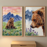 Two rectangular framed artworks hang side-by-side on a neutral wall. Each features a brown bear in a serene natural setting, rendered in a soft watercolor style. The piece on the left shows a full-bodied bear sitting in a vibrant green meadow dotted with small, colorful wildflowers, set before jagged blue mountains under a warm pink and orange sky. The piece on the right displays a close-up profile of a bear's head, gazing down at a patch of purple and blue flowers, with faint blue mountains.
