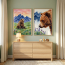 Two large, framed watercolor illustrations featuring brown bears are prominently displayed on a light sage green wall. The illustration on the left depicts a bear sitting in a grassy field filled with wildflowers, with a backdrop of jagged blue mountains under a soft pink and orange sky. The illustration on the right is a detailed close-up of a bear's face, looking towards a cluster of purple flowers, with mountains faintly visible in the background.
