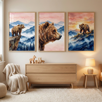 A set of three framed watercolor-style illustrations featuring brown bears hangs on a beige wall in a cozy room. The central piece is a close-up of a bear's head, while the flanking scenes depict bears in a misty, blue mountain landscape under a pink and orange sunset sky. Below the wall art, a light wood dresser holds small wooden animal toys. To the left, a comfortable chair is draped with a textured knit blanket, and to the right, a glowing lamp sits on a small wooden side table.