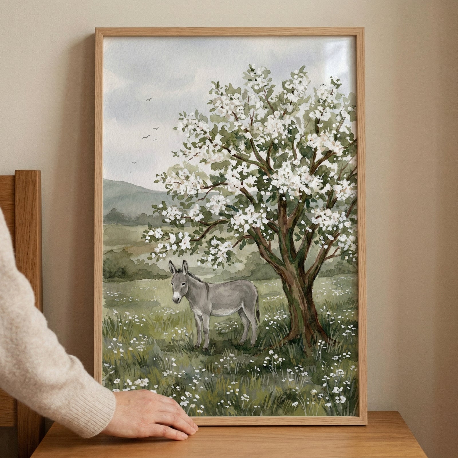 A framed watercolor-style illustration depicts a peaceful countryside scene. A grey donkey stands in a lush green meadow filled with small white wildflowers. A large, blossoming tree with many delicate white flowers shades the donkey. 