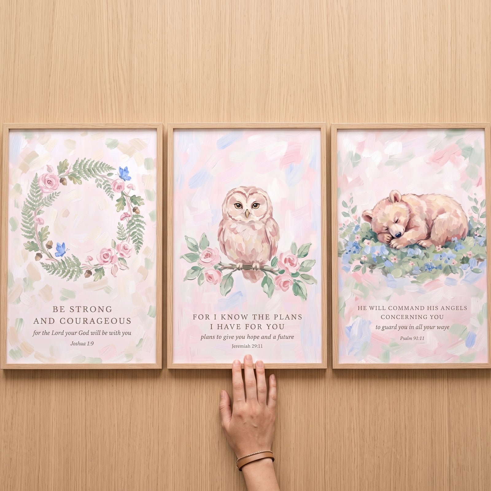 A hand touches a watercolor blush floral scripture bible wall art set of 3 featuring a wreath, owl, and sleeping bear cub.