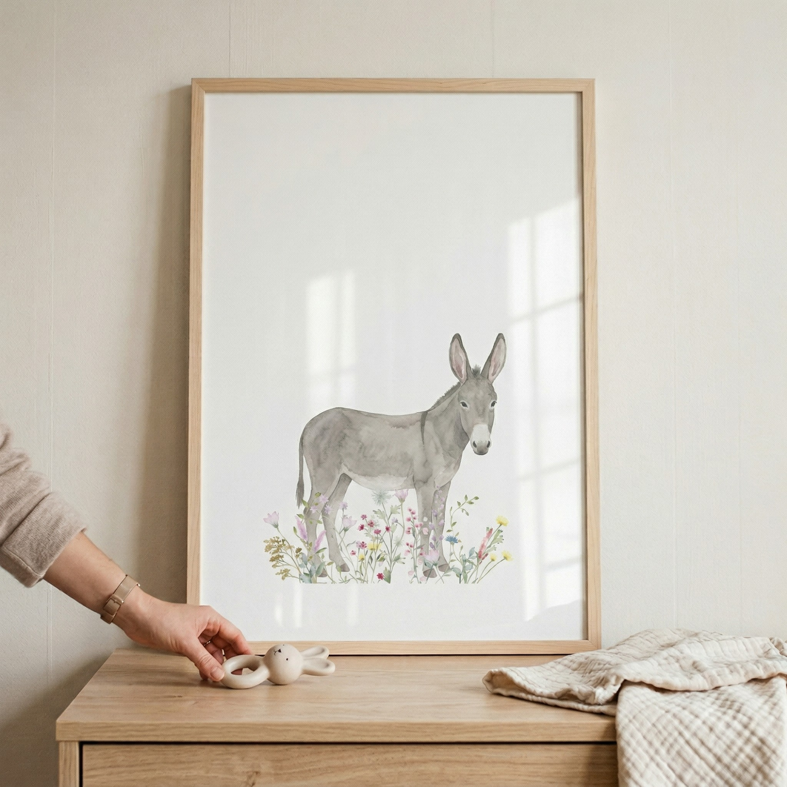 A watercolor-style illustration of a gray donkey standing in a patch of delicate, colorful wildflowers in shades of pink, purple, and yellow. The art is set in a light wood frame, which leans against an off-white wall on a wooden surface. To the left, a person's hand reaches toward a small wooden toy animal. A soft, textured cream-colored cloth is draped on the right, creating a warm and gentle nursery-like scene.