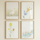 Four framed watercolor illustrations of white ducks are displayed in a square grid on a neutral wall. Each piece is set in a light-toned wood frame. The top-left art shows a duck walking through yellow wildflowers. The top-right image is a close-up of a duck's head looking at a blue flower. On the bottom-left, a duck peeks from behind a tall stalk of yellow blossoms. The bottom-right piece features a duck by a blue stream with small flowers. The style is soft, minimalist, and whimsical.