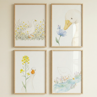 Four framed watercolor illustrations of white ducks are displayed in a square grid on a neutral wall. Each piece is set in a light-toned wood frame. The top-left art shows a duck walking through yellow wildflowers. The top-right image is a close-up of a duck's head looking at a blue flower. On the bottom-left, a duck peeks from behind a tall stalk of yellow blossoms. The bottom-right piece features a duck by a blue stream with small flowers. The style is soft, minimalist, and whimsical.