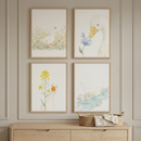 Four framed watercolor-style illustrations are arranged in a grid on a light-colored, paneled wall. Each piece features a white duck in a soft, natural scene. The top-left shows a duck in a field of yellow flowers; the top-right shows a duck head near a blue flower; the bottom-left shows a duck by a tall yellow flower; and the bottom-right shows a duck by a stream. The art is in light wood frames above a matching wooden dresser. A small woven basket with a cloth sits on the dresser top.