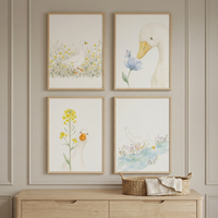 Four framed watercolor-style illustrations are arranged in a grid on a light-colored, paneled wall. Each piece features a white duck in a soft, natural scene. The top-left shows a duck in a field of yellow flowers; the top-right shows a duck head near a blue flower; the bottom-left shows a duck by a tall yellow flower; and the bottom-right shows a duck by a stream. The art is in light wood frames above a matching wooden dresser. A small woven basket with a cloth sits on the dresser top.