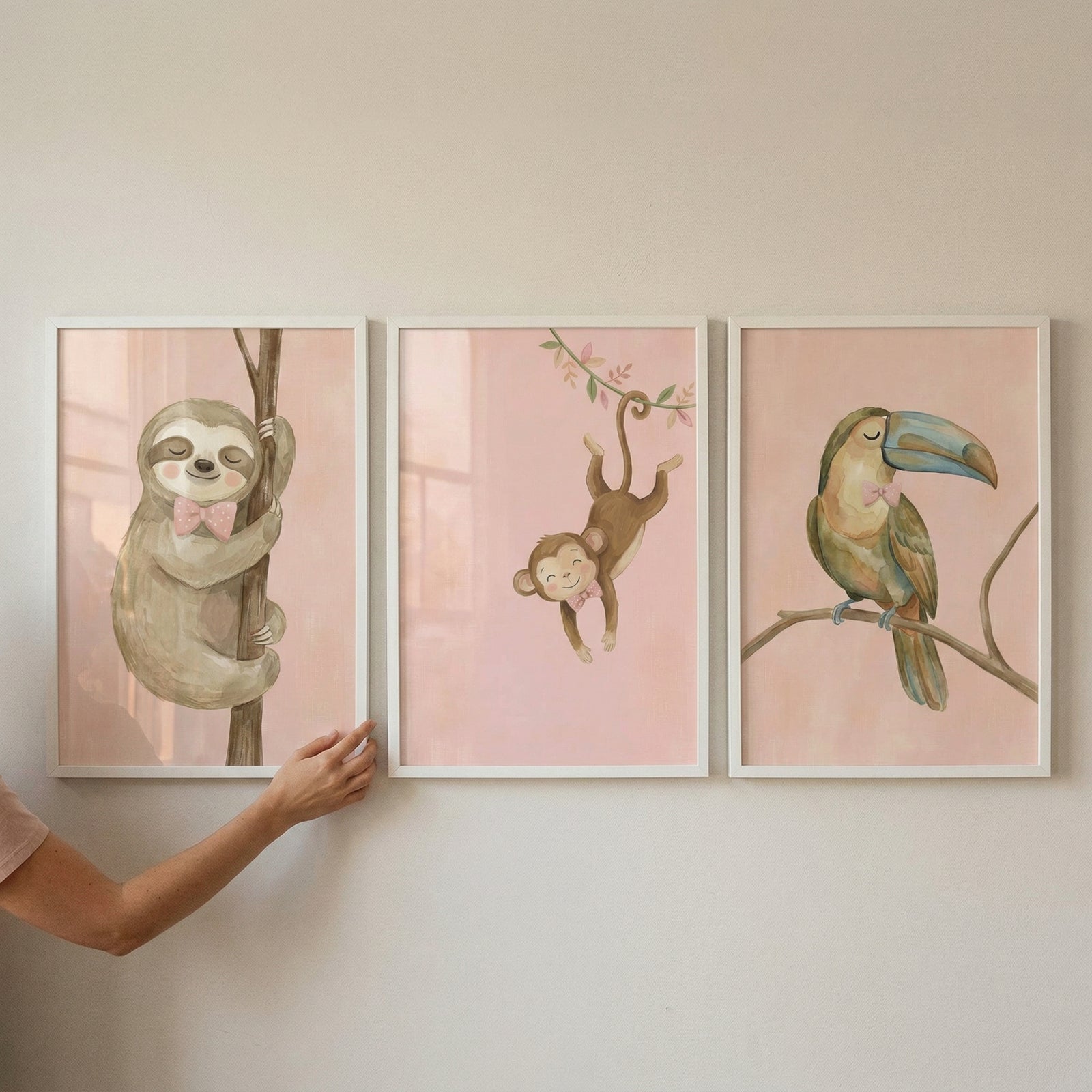Three white-framed watercolor illustrations are arranged in a horizontal row on a light-colored wall. Each artwork depicts a cute jungle animal on a soft pink background, with every animal wearing a small pink bow tie. 
