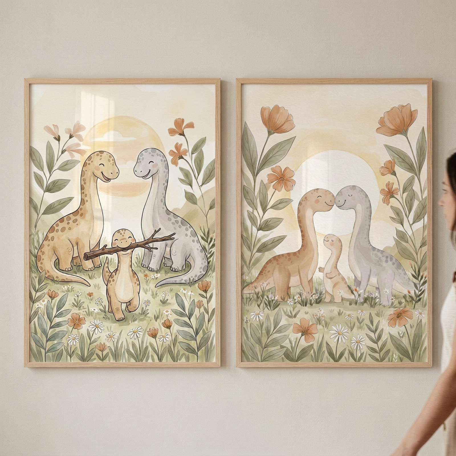 Two framed watercolor-style illustrations hang side-by-side on a plain wall. The left print depicts two large, long-necked dinosaurs—one tan with spots and one light blue—looking down at a smaller tan dinosaur holding a stick in its mouth. The right print features the same adult dinosaurs touching noses, with the small dinosaur standing between them. Both scenes are set in a meadow of tall grass and wildflowers under a large sun, rendered in soft, muted tones of beige, green, and orange.