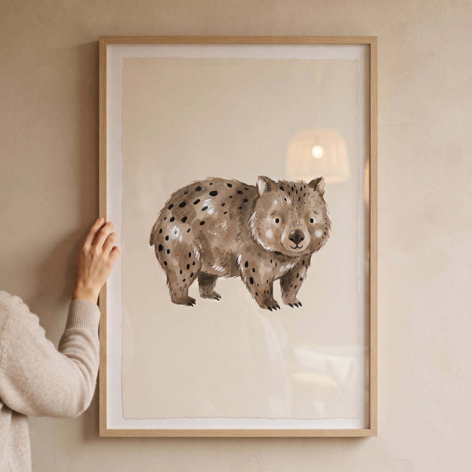 Watercolor brown spotted wombat wall art in a light wood frame held against a minimalist beige wall.