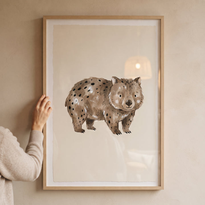 Watercolor brown spotted wombat wall art in a light wood frame held against a minimalist beige wall.
