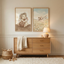 A cozy, neutral-toned nursery features a light wood dresser with six drawers. A white fringed throw is draped over one side. On top, a small ceramic lamp glows warmly next to simple wooden decor. Above the dresser hang two framed illustrations of a cartoon rabbit in a bonnet; one shows the rabbit with yarn, the other running through a meadow. A woven basket with wooden toys sits on the floor beside a white shag rug. The walls have cream-colored paneling under soft, natural light.