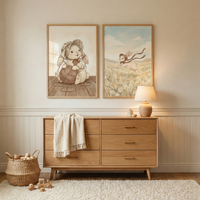 A cozy, neutral-toned nursery features a light wood dresser with six drawers. A white fringed throw is draped over one side. On top, a small ceramic lamp glows warmly next to simple wooden decor. Above the dresser hang two framed illustrations of a cartoon rabbit in a bonnet; one shows the rabbit with yarn, the other running through a meadow. A woven basket with wooden toys sits on the floor beside a white shag rug. The walls have cream-colored paneling under soft, natural light.
