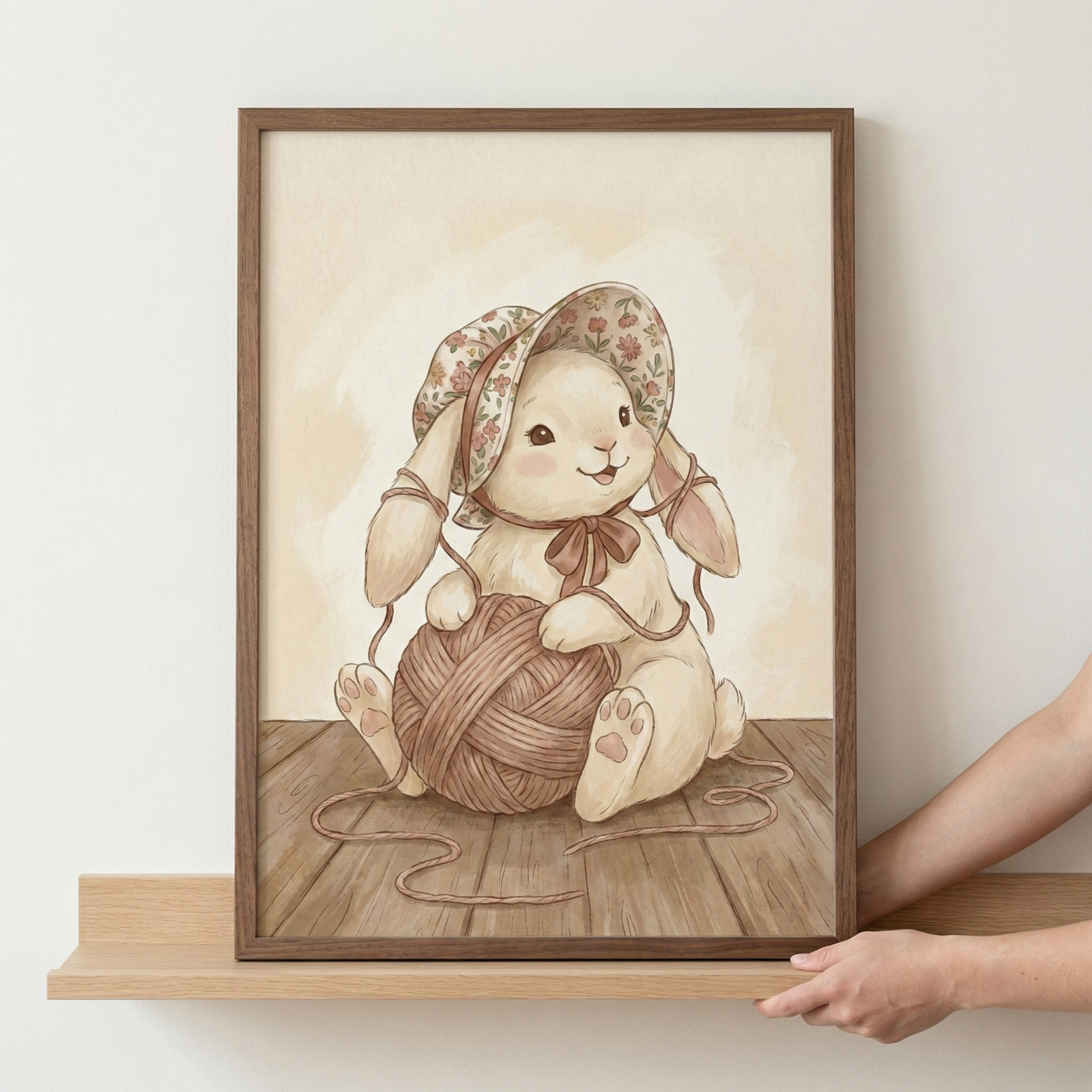 A person's hands hold a wooden-framed illustration of a sweet, cream-colored bunny rabbit. The bunny sits on a wooden floor, cradling a large ball of brown yarn. It wears a floppy sun hat with a red floral print, tied with a ribbon. The artwork has a soft, watercolor-like aesthetic in warm, neutral tones. The frame rests on a light wood shelf against a plain white wall.