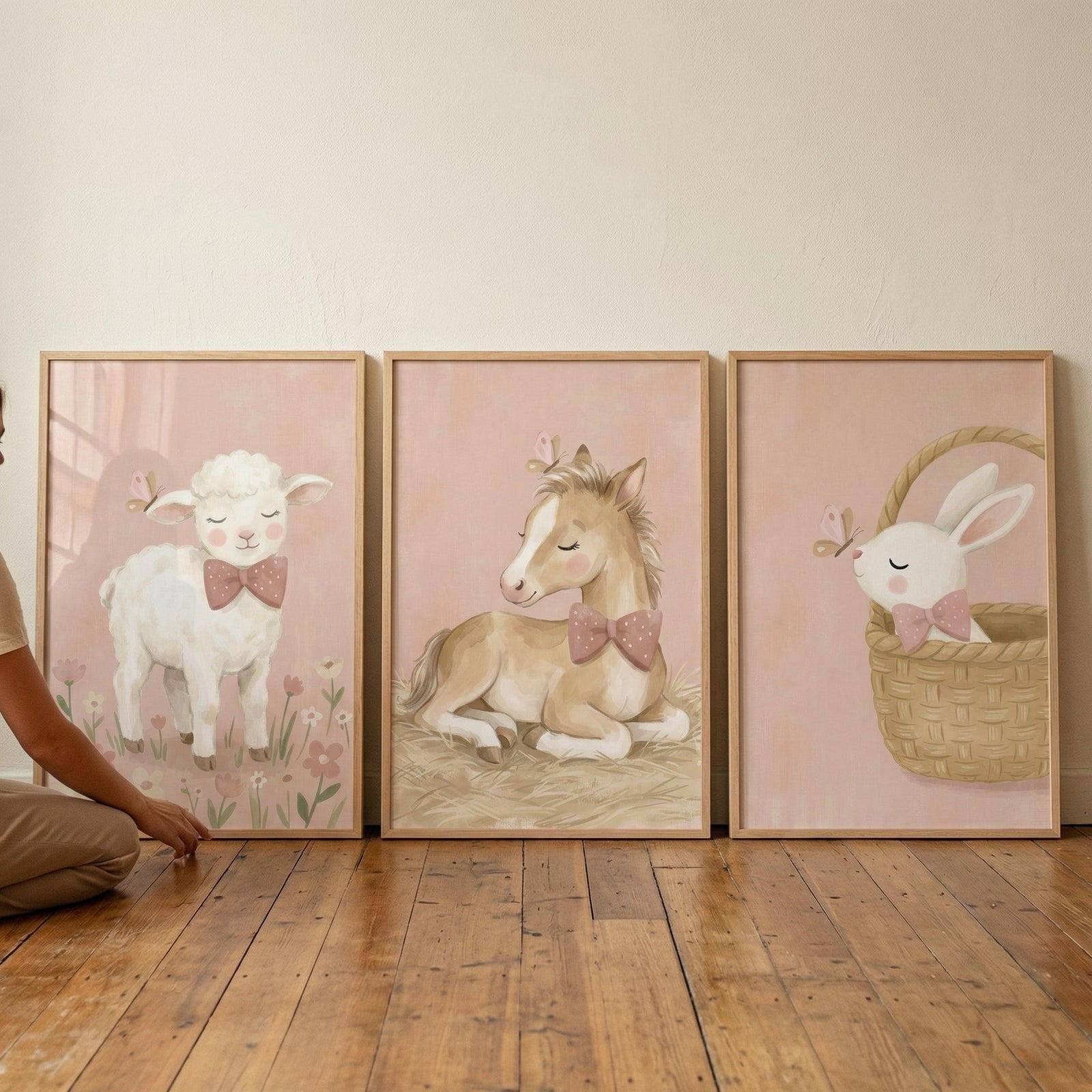 An image displays three large, vertically-oriented wooden frames leaning against a cream-colored wall on a rustic wooden floor. Each frame contains a whimsical illustration of a baby animal on a soft pink background. On the left, a fluffy white lamb stands in a small patch of flowers wearing a pink bow tie. 