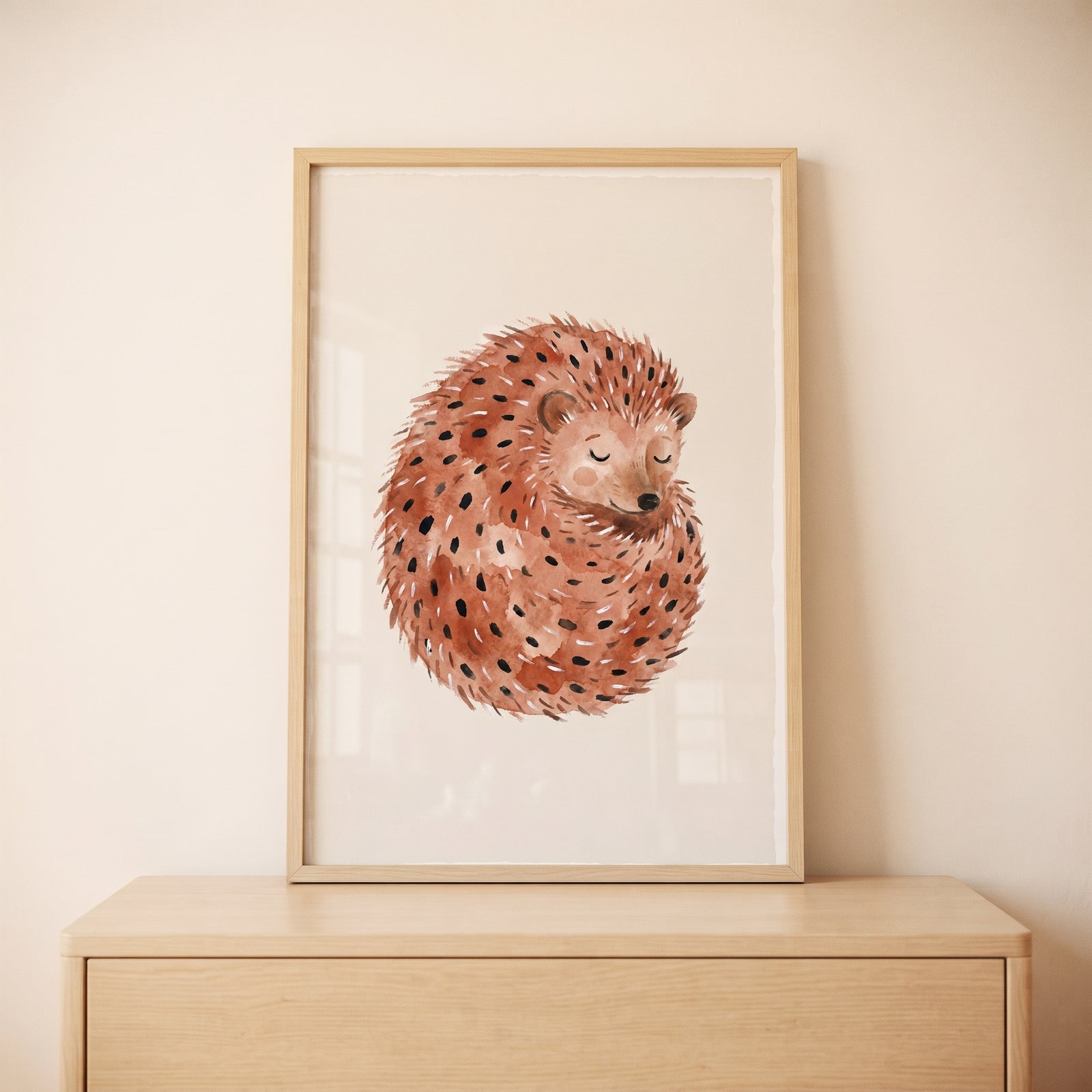 Watercolor curled up hedgehog wall art in a wood frame resting on a light wood dresser against a neutral wall.