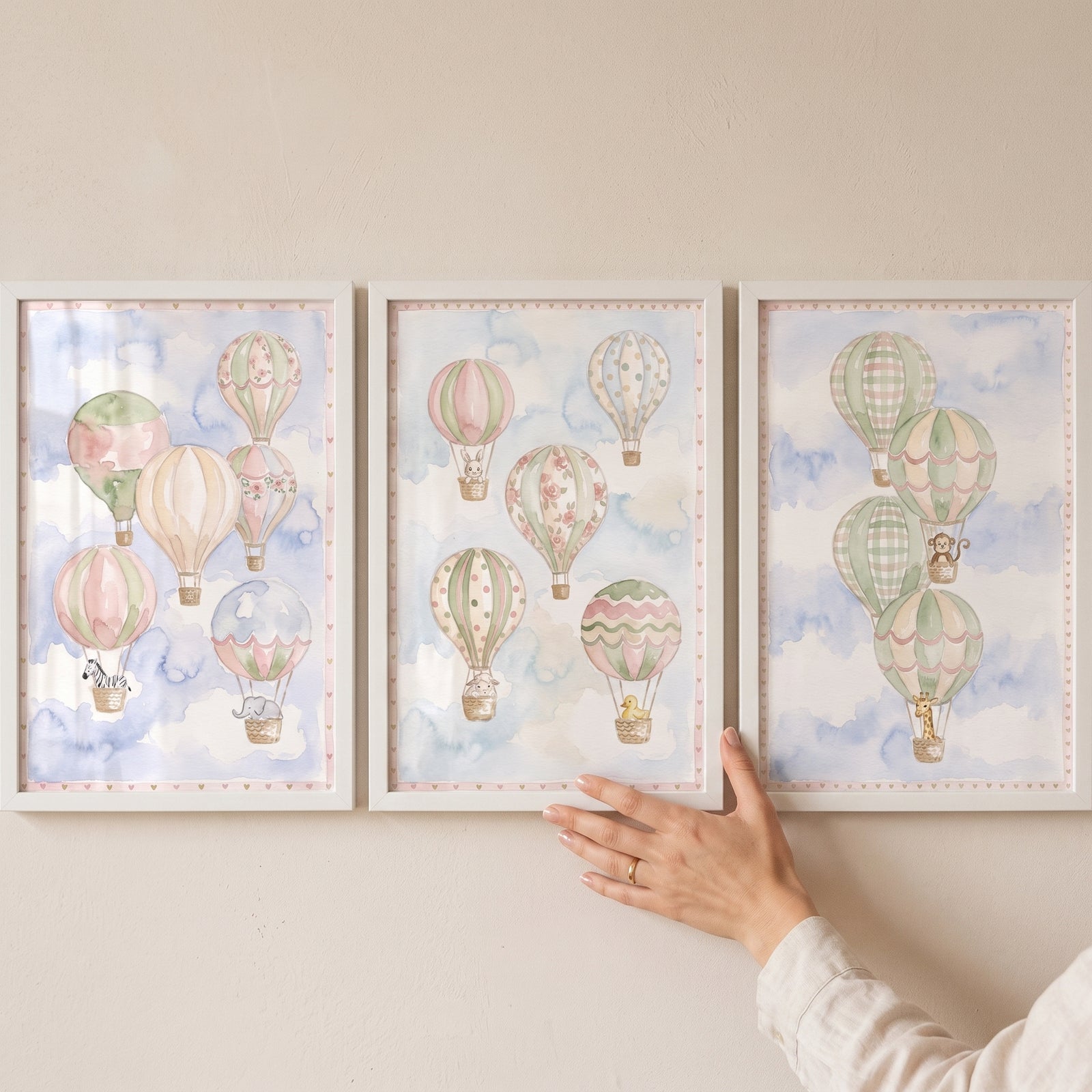 A watercolor dreamy animal balloon wall art set of 3 hangs on a wall as a hand adjusts the middle framed print.