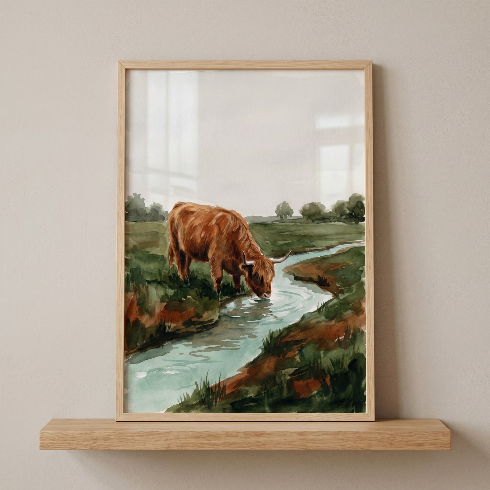 Framed watercolor drinking highland cow wall art on a shelf shows a brown cow by a stream in a green field.