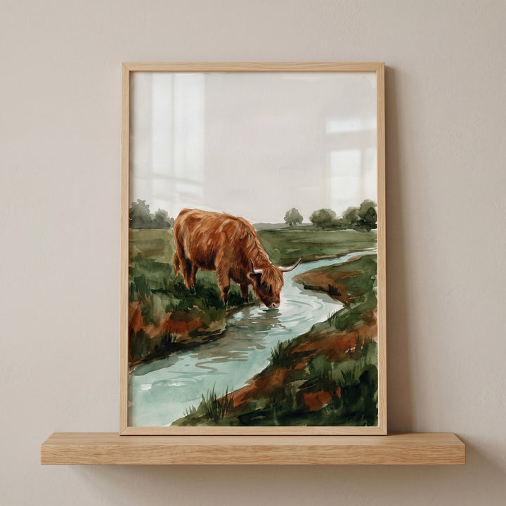 Framed watercolor drinking highland cow wall art on a shelf shows a brown cow by a stream in a green field.