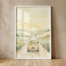 A vertical framed watercolor illustration leans against a plain, neutral wall on a light wood floor. The artwork depicts a whimsical scene with a small, rounded yellow vintage car driving away down a long dirt road towards a bright sun setting behind soft, rolling green hills. 