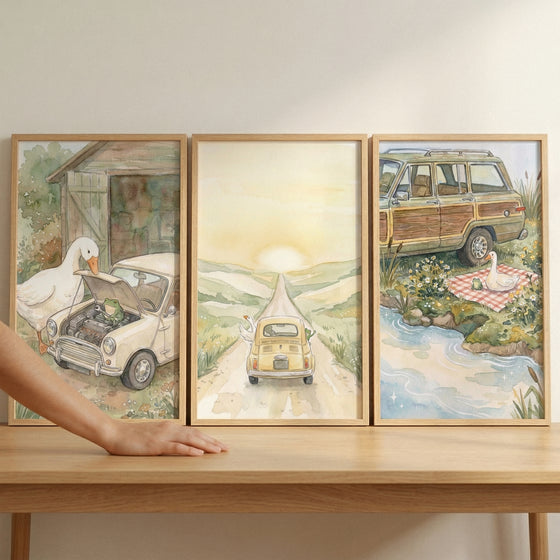 Three rectangular wooden-framed pieces of art are displayed side-by-side on a light-colored wooden surface. The art features whimsical, soft-toned watercolor illustrations centered around vintage vehicles and white geese in pastoral settings.