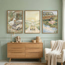 A cozy room features soft sage green walls with elegant decorative molding. Three framed watercolor-style illustrations are hung side-by-side above a light wood dresser. 
