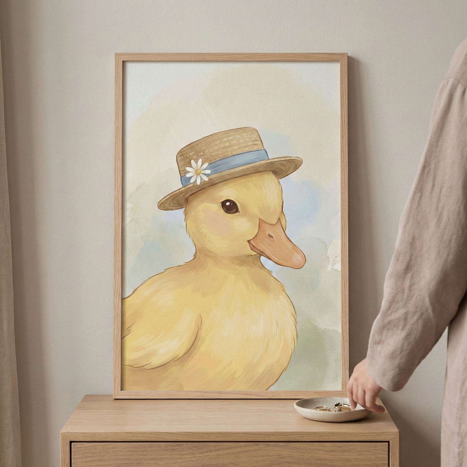 A framed illustration features a charming yellow duckling in a portrait pose. The duckling wears a small straw hat embellished with a light blue ribbon and a single white daisy. The artwork has a soft, whimsical style with visible brushwork and a gentle pastel background of blue and cream tones. The light wood frame rests on a wooden dresser against a plain, neutral wall. To the right, a person's hand reaches toward a small white ceramic dish containing rings, situated at the base of the frame.