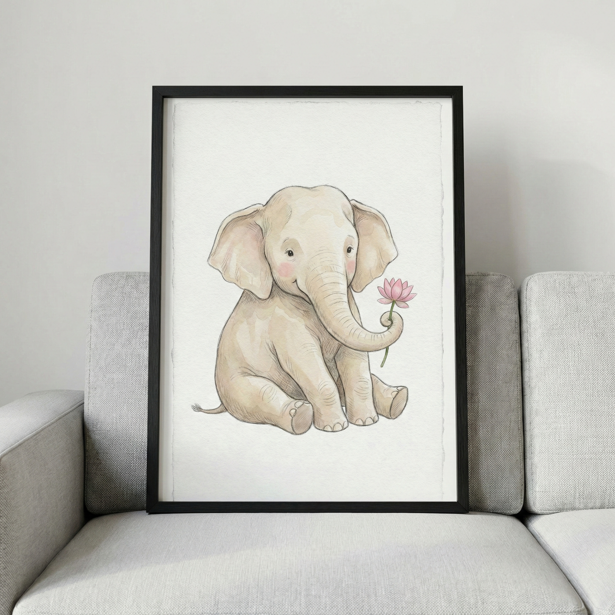 A vertically oriented, black-framed illustration of a young elephant rests on a light gray fabric sofa against a plain off-white wall. The artwork features a soft, beige-colored cartoon elephant sitting down with a gentle expression and rosy pink cheeks. Its trunk is curled upward, holding a single pink flower with a green stem. The illustration is created with a soft, delicate style on a textured off-white background.
