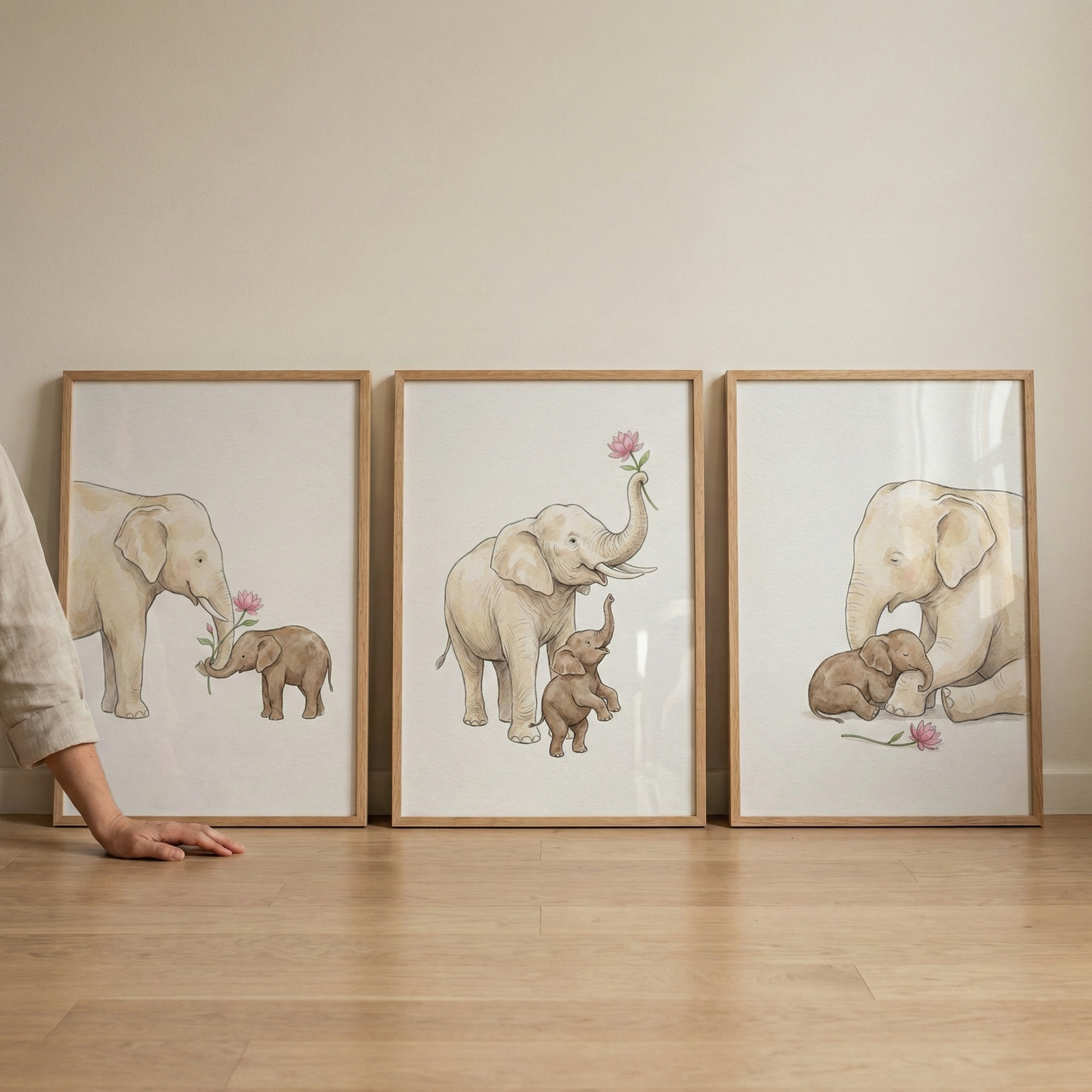 Three wooden-framed illustrations of elephants lean against a light-colored wall on a wooden floor. Each artwork depicts an adult elephant and a small calf in soft, muted tones. In the first, the adult offers a pink flower to the calf. In the second, the adult holds the flower high while the calf reaches up. In the third, the adult elephant sits protectively beside the sleeping calf with the pink flower nearby. A person's hand rests on the floor next to the left-most frame.