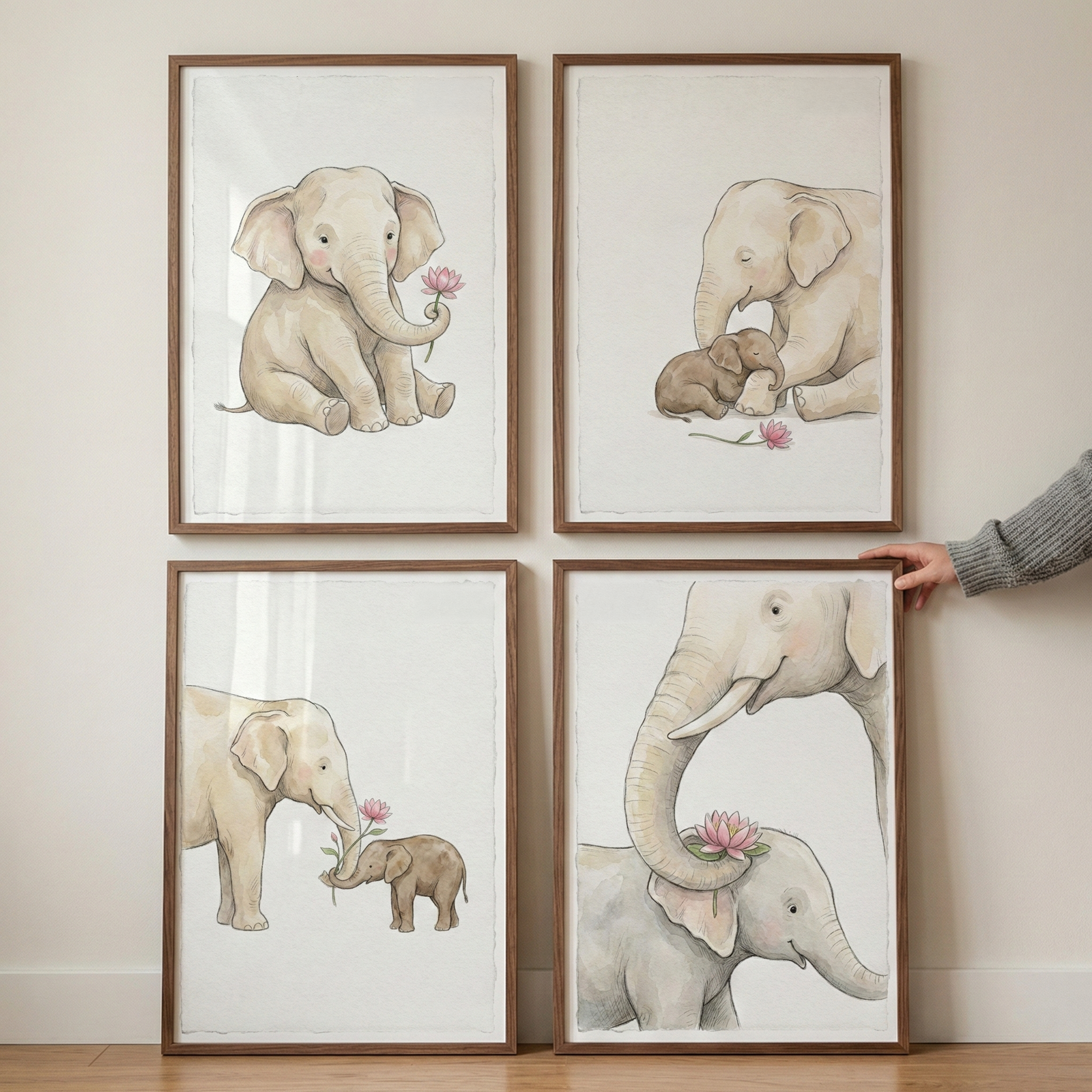 Four wooden frames containing soft-toned elephant illustrations are mounted on a plain wall in a square arrangement. The watercolor-style images feature various scenes: a single elephant calf with a flower, an adult elephant with a baby elephant, and close-up interactions between the animals and pink blossoms. The color palette consists of neutral beiges and light pinks. A person's hand is visible on the right side, touching the frame on the bottom right.