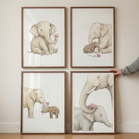 Four wooden frames containing soft-toned elephant illustrations are mounted on a plain wall in a square arrangement. The watercolor-style images feature various scenes: a single elephant calf with a flower, an adult elephant with a baby elephant, and close-up interactions between the animals and pink blossoms. The color palette consists of neutral beiges and light pinks. A person's hand is visible on the right side, touching the frame on the bottom right.