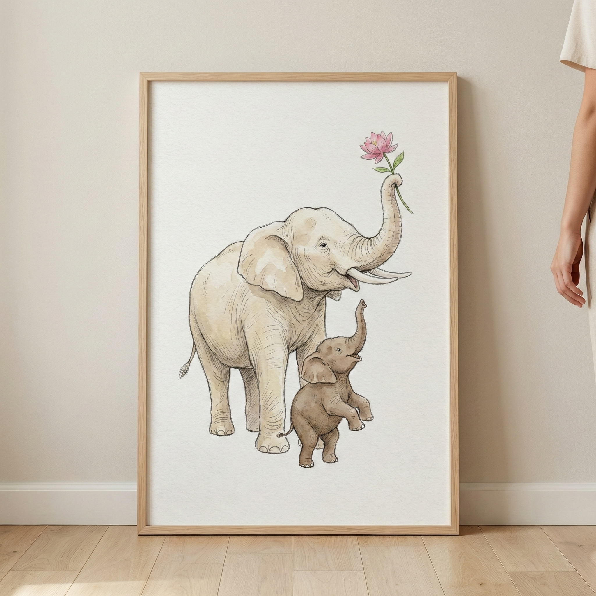 A framed art piece leans against a neutral wall on a light wood floor. The simple wooden frame holds an illustration of a large cream elephant and a smaller brown calf. The adult elephant raises its trunk to hold a single pink flower, while the baby elephant stands on its hind legs, reaching up toward it. The artwork features soft colors and a watercolor-like style on a textured background. To the right, a person's arm and the edge of a light-colored garment are partially visible.
