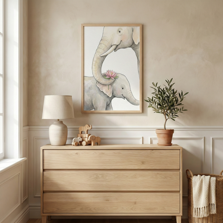 A serene nursery interior featuring a vertical framed illustration on a beige wall. The artwork depicts a soft-toned adult elephant gently placing a pink flower on a baby elephant's head using its trunk. Below the frame, a light wood dresser with three drawers holds a small lamp with a cream base, a set of wooden elephant figurines, and a small plant in a terracotta pot. A woven basket with a fringed blanket sits nearby. The room is finished with classic white wainscoting and soft light.