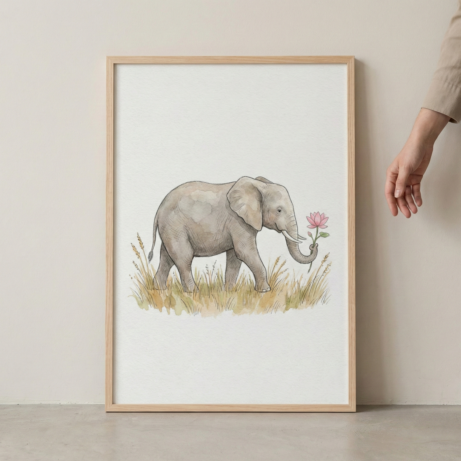 A vertically oriented watercolor illustration presented in a light-colored wooden frame. The artwork features a gentle gray elephant walking through a field of tall, golden-brown grasses. The elephant is shown in profile and holds a delicate pink flower in the tip of its trunk. The background is a clean, off-white color. The frame rests against a neutral-colored wall, and a human hand is visible on the right side of the frame.