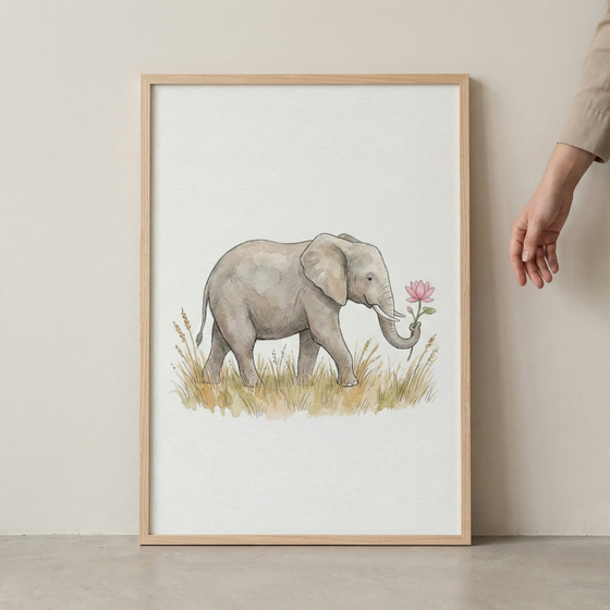 A vertically oriented watercolor illustration presented in a light-colored wooden frame. The artwork features a gentle gray elephant walking through a field of tall, golden-brown grasses. The elephant is shown in profile and holds a delicate pink flower in the tip of its trunk. The background is a clean, off-white color. The frame rests against a neutral-colored wall, and a human hand is visible on the right side of the frame.