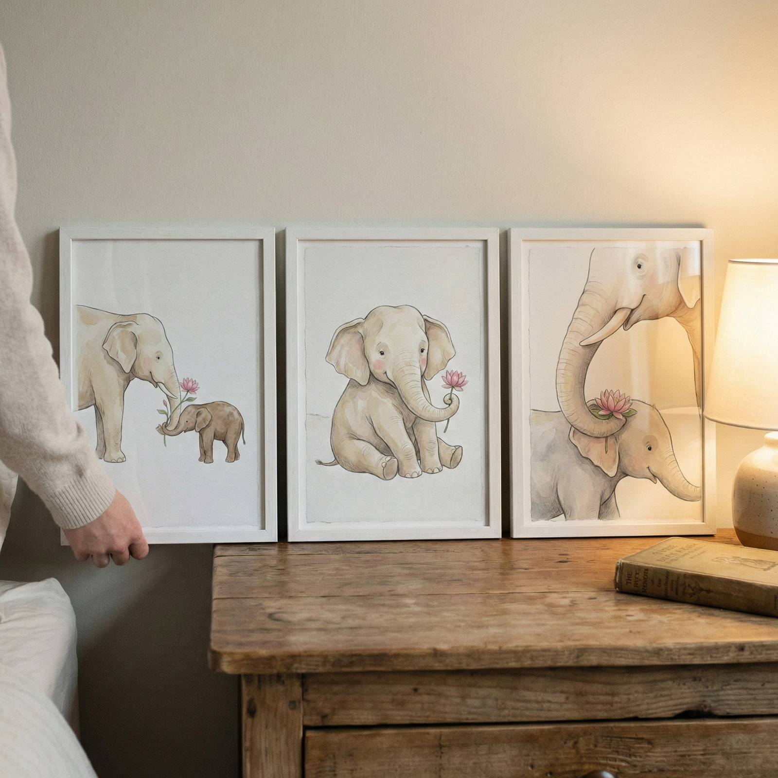 Three framed watercolor-style illustrations of cute elephants rest on a rustic wooden tabletop. A hand holds the frame on the left, which shows an adult elephant offering a pink flower to a smaller baby elephant. The center frame features a sitting baby elephant holding a pink lotus blossom. The frame on the right captures an adult elephant’s trunk gently resting on a baby elephant's head. A soft glow from a nearby lamp illuminates the scene alongside an old book on the wooden surface.