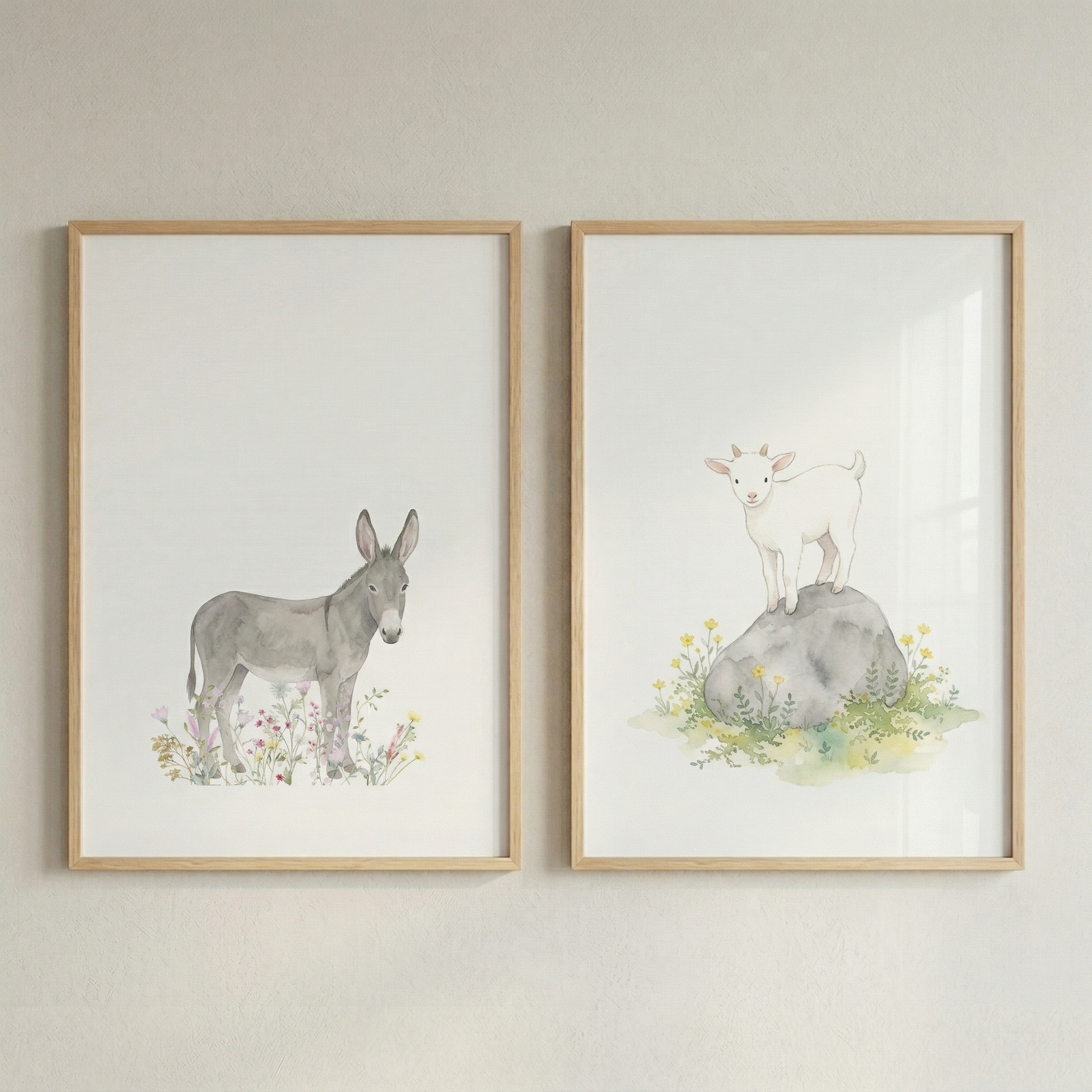 Two vertical light wood frames hang side-by-side on a neutral wall. Each frame holds a watercolor-style illustration of a baby animal on a white background. The frame on the left depicts a grey donkey foal standing in a bed of pastel-colored wildflowers. The frame on the right depicts a small white goat standing atop a smooth grey stone, which is nestled among green grass and yellow blossoms. The artwork has a soft, whimsical feel, suitable for a nursery.
