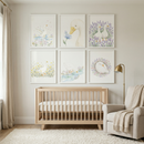 A neutral-toned nursery features a light wood six-drawer dresser against a cream wall. Above the dresser hang three framed illustrations: a white waterfowl in a floral wreath, a duck looking at a blue flower, and two ducks in a lavender field. On the dresser are a small lamp, a woven basket, and folded linens. A beige curtain hangs on the left, and a potted olive tree stands on the right. The floor is covered by a light-colored woven rug, creating a serene and rustic atmosphere.