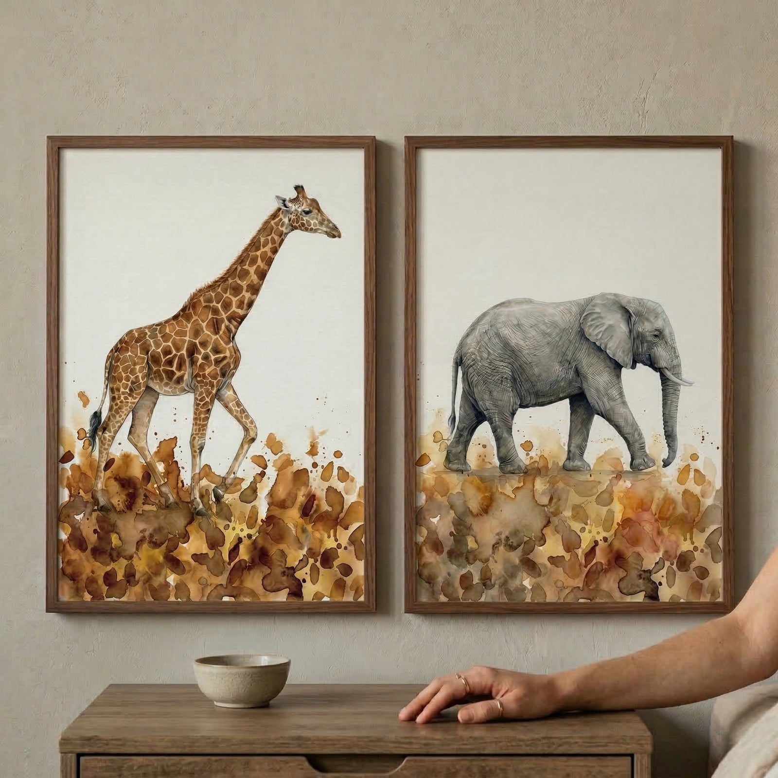 This image features a set of two vertical art pieces in thin, dark wood frames, hung side-by-side on a light beige textured wall. The art on the left depicts a giraffe in a detailed watercolor style, rendered in warm brown tones. The art on the right features an elephant, also in a watercolor style, using varying shades of grey. Both animals are set against a plain white background, with a base of abstract, earth-toned watercolor splatters at their feet. 
