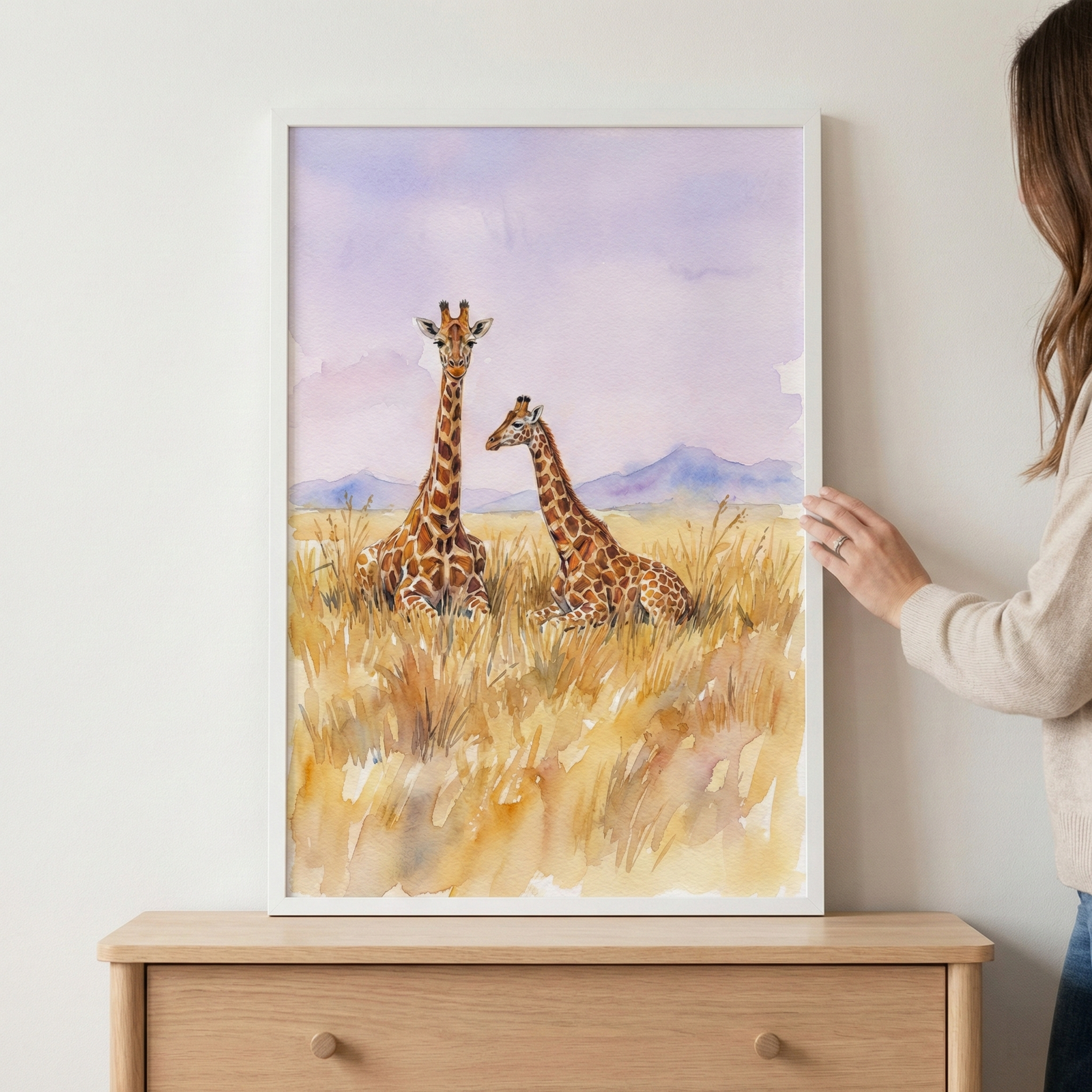 A vertical watercolor illustration of two giraffes resting in a field of tall, golden-brown grass is displayed in a thin white frame. The larger giraffe faces forward, while a smaller one beside it looks to the side. Behind them, soft purple washes create a sky above distant blue mountains. The framed piece sits atop a natural wood dresser against a pale wall. A person's hand with a ring is visible on the right side, gently holding the edge of the frame.