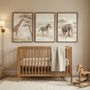 A serene nursery features a light wood crib centered against a warm beige wall. Above the crib, three framed watercolor-style illustrations depict African animals in muted, earthy tones: a giraffe near a tree, a galloping zebra, and a walking elephant. To the left, a simple wall-mounted sconce with a white shade adds a soft glow. A cream-colored blanket is draped over the crib's rail. On the floor, a plush off-white rug holds a minimalist wooden rocking horse to the right of the crib.