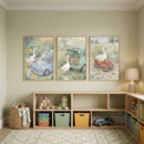 A serene and light-filled child's playroom features a row of three framed watercolor-style illustrations hung above a low wooden storage unit. The walls are painted a soft sage green, creating a peaceful atmosphere.
