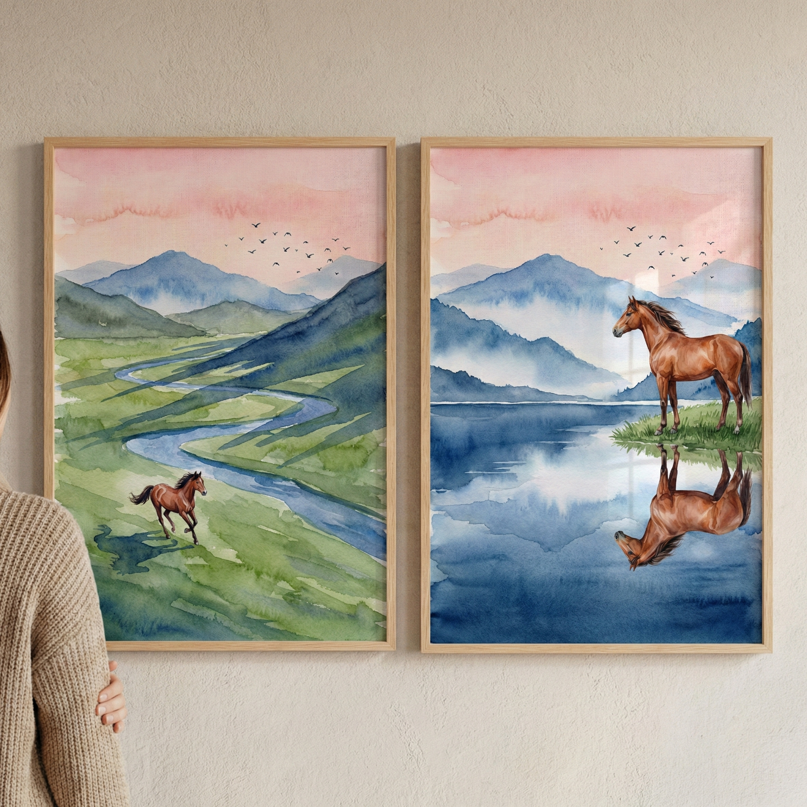 A vertical image features two side-by-side framed watercolor-style illustrations hanging on a textured, off-white wall. On the left, a person wearing a beige sweater is partially visible. The left illustration shows a brown horse running through a lush green landscape with a winding blue river, set against soft blue mountains under a pink and orange sky. The right illustration depicts a larger brown horse standing at the edge of a blue lake, with its reflection clear in the water, backed by similar mountain