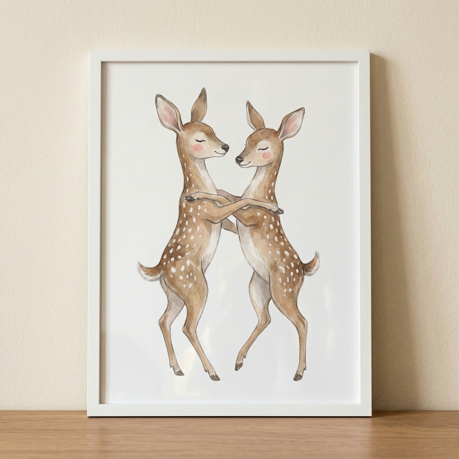 Watercolor hugging spotted fawn wall art shows two fawns embracing in a white frame on a wooden table.